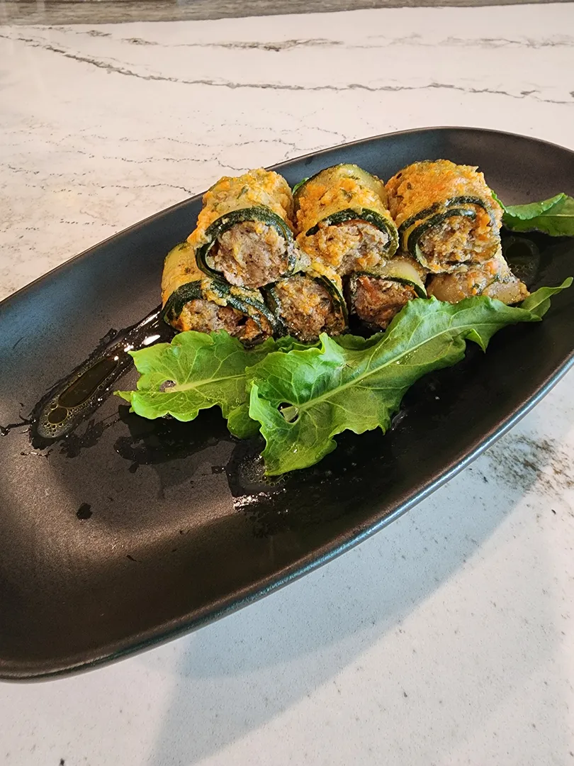 Four zucchini roll-ups filled with a meat mixture, served on a black plate with a side of leafy greens and a drizzle of oil.