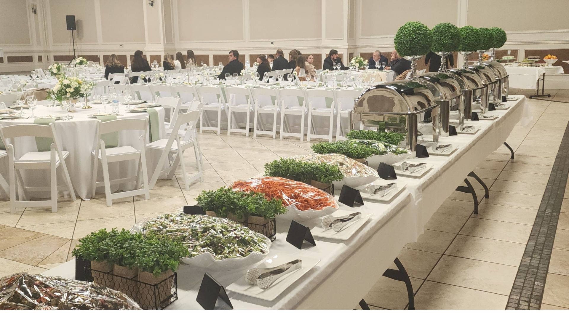A long buffet table with covered trays and salads is set up in front of round dining tables with guests seated at an indoor event venue.