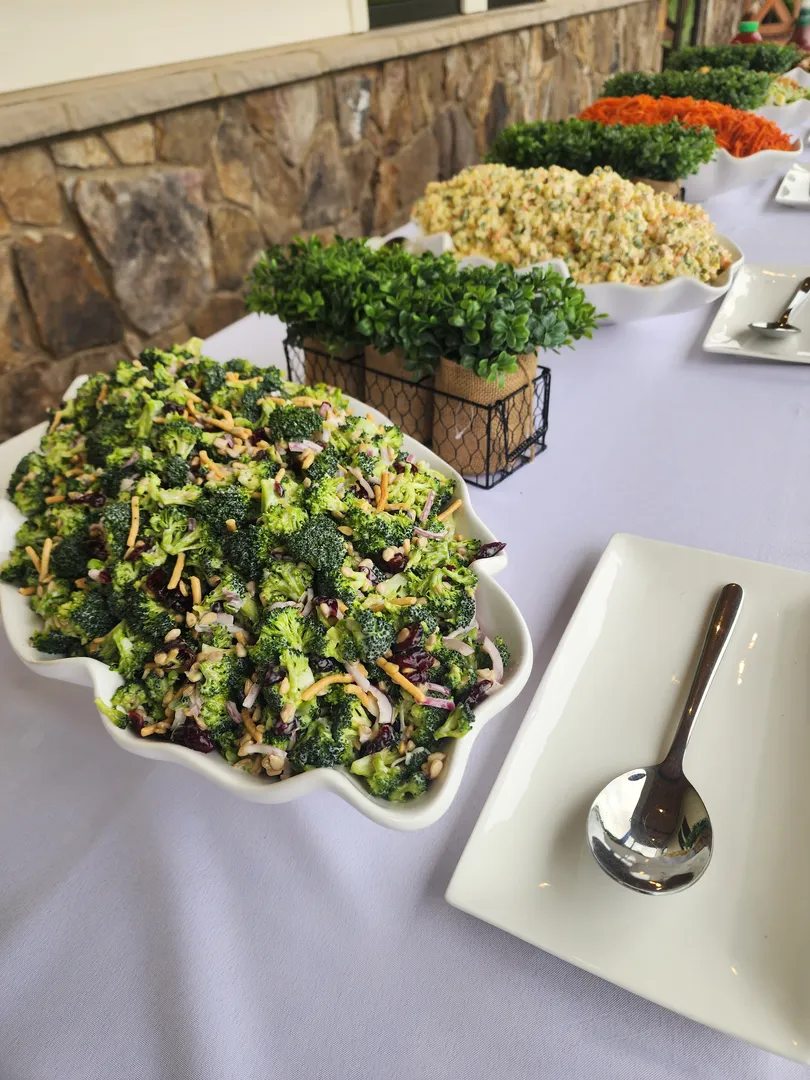 A buffet table with dishes of salads, including broccoli salad in the foreground, and trays of fresh microgreens on a white tablecloth.