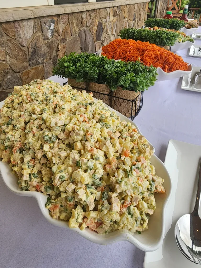 A large platter of mixed vegetable salad with creamy dressing is displayed on a buffet table, with shredded carrots and potted plants in the background.