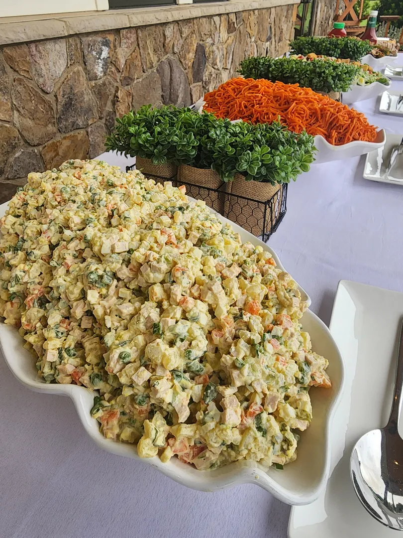 A large platter of creamy potato salad sits on a buffet table next to decorative green plants and another platter filled with shredded carrots.