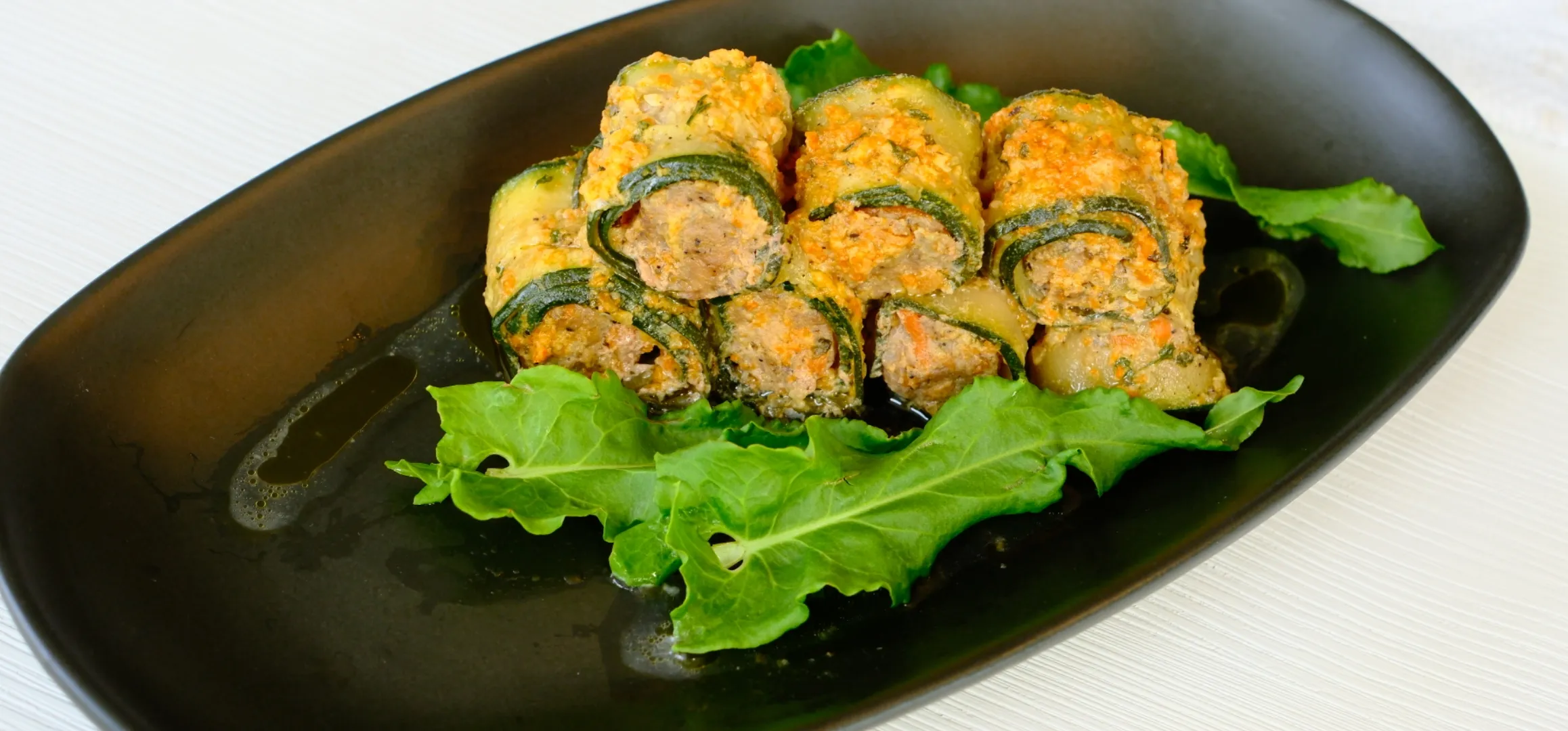 Four zucchini roll-ups stuffed with a breadcrumb filling are arranged on a black plate, garnished with fresh lettuce leaves.