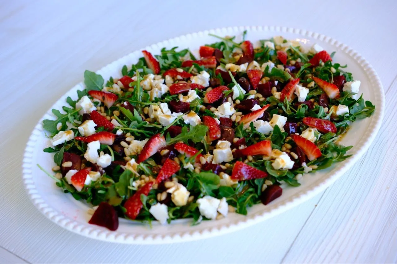 A white oval platter with arugula salad topped with sliced strawberries, crumbled feta cheese, walnuts, and what appear to be chopped beets.