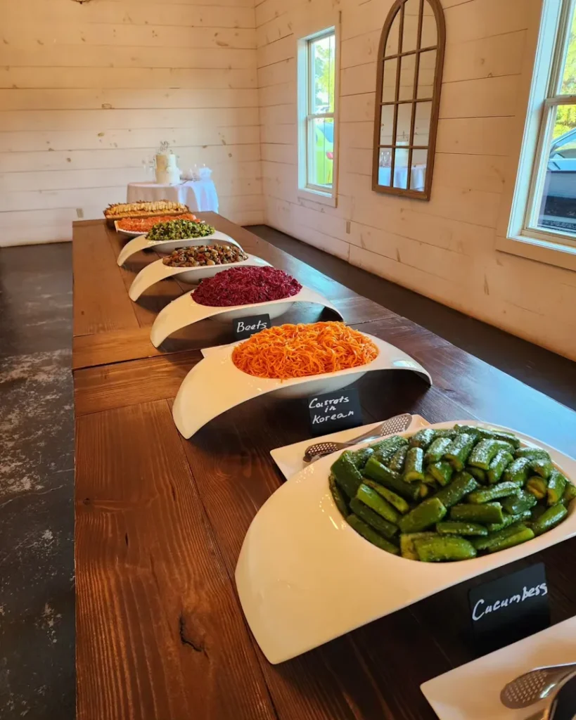 A row of white bowls filled with various salads and vegetables is arranged on a wooden table, each labeled with handwritten signs such as "Cucumbers" and "Carrots Korean.