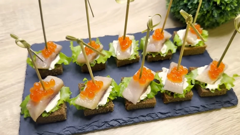A slate platter with ten bite-sized appetizers, each with dark bread, lettuce, cream, a slice of fish, orange caviar, and a skewer for serving.